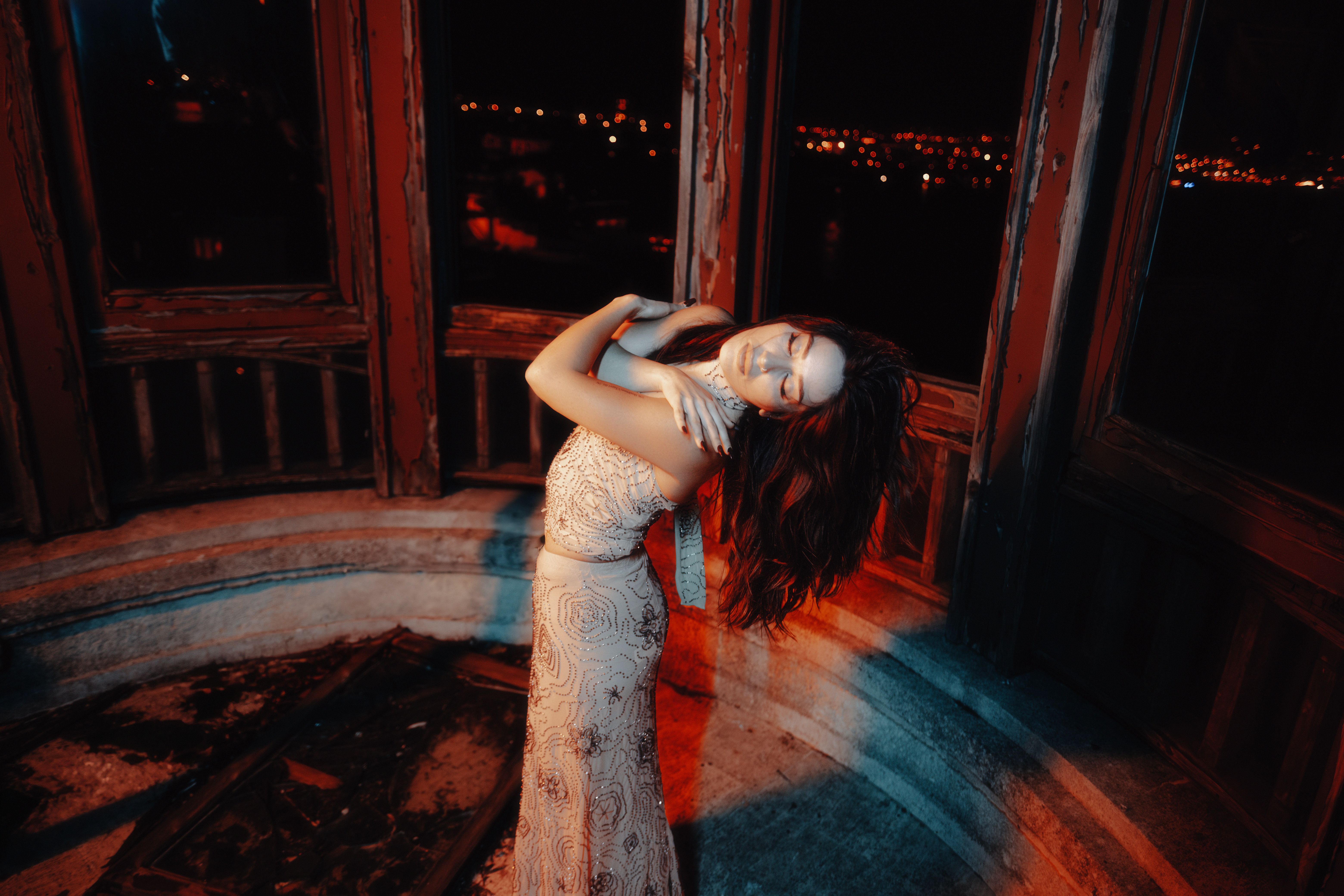 Woman in a decorative gown posing indoors at night with city lights in the background.