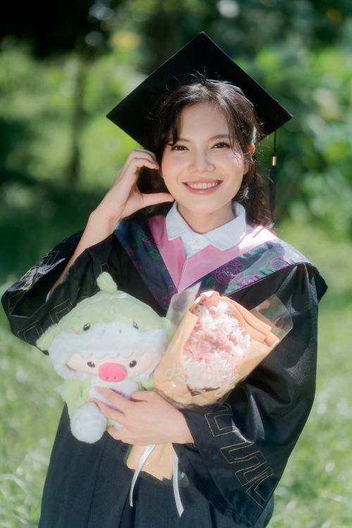 Free Young adult woman in graduation gown holds plush toy and flowers, celebrating achievement outdoors. Stock Photo