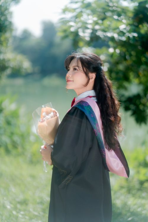 Free Graduate holding a bouquet and smiling in a serene outdoor setting. Stock Photo