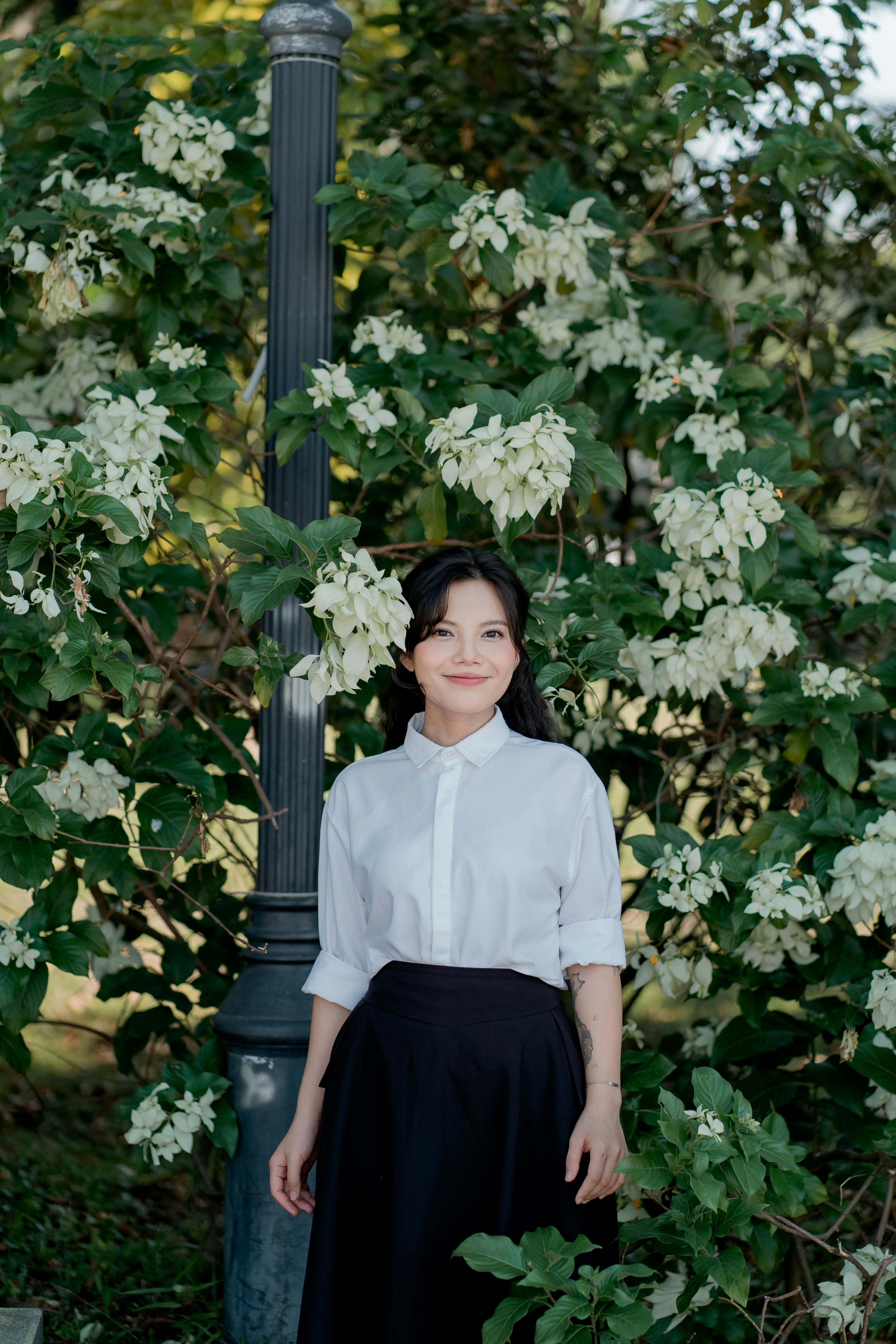 Woman in white blouse posing naturally by a lamppost and white flowers outdoors.