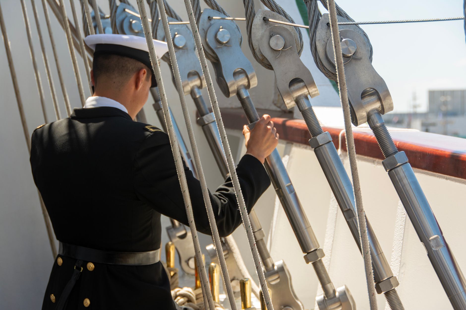 A sailor in uniform adjusts ropes on a ship deck during the day.