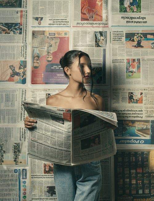 Free A woman reads a newspaper against a wall covered in newsprints, creating an artistic and moody atmosphere. Stock Photo