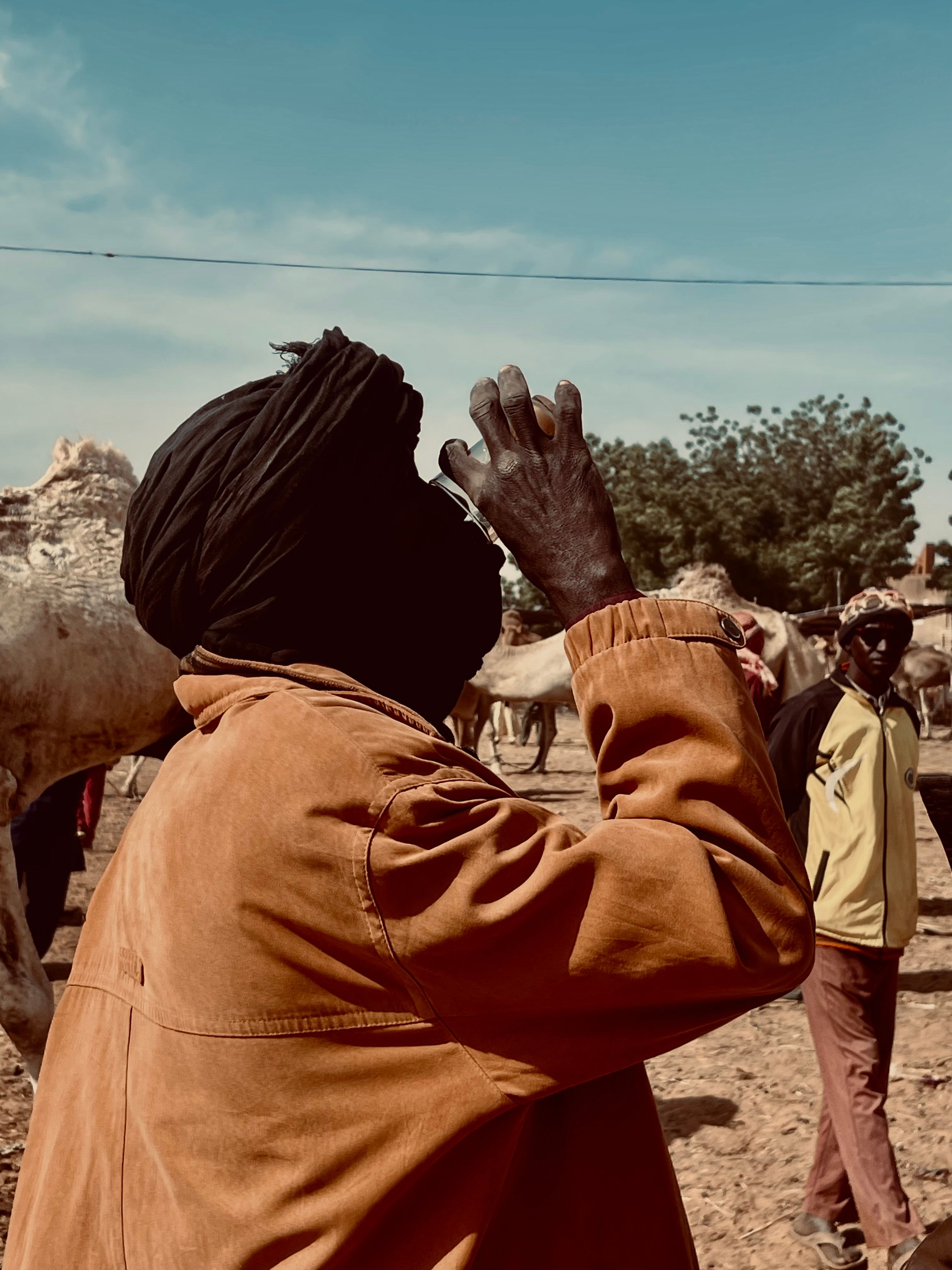 A camel herder in a traditional turban at an outdoor livestock market under a bright sky.
