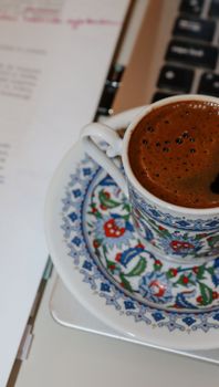 A cup of rich Turkish coffee in a decorated cup placed beside a laptop.