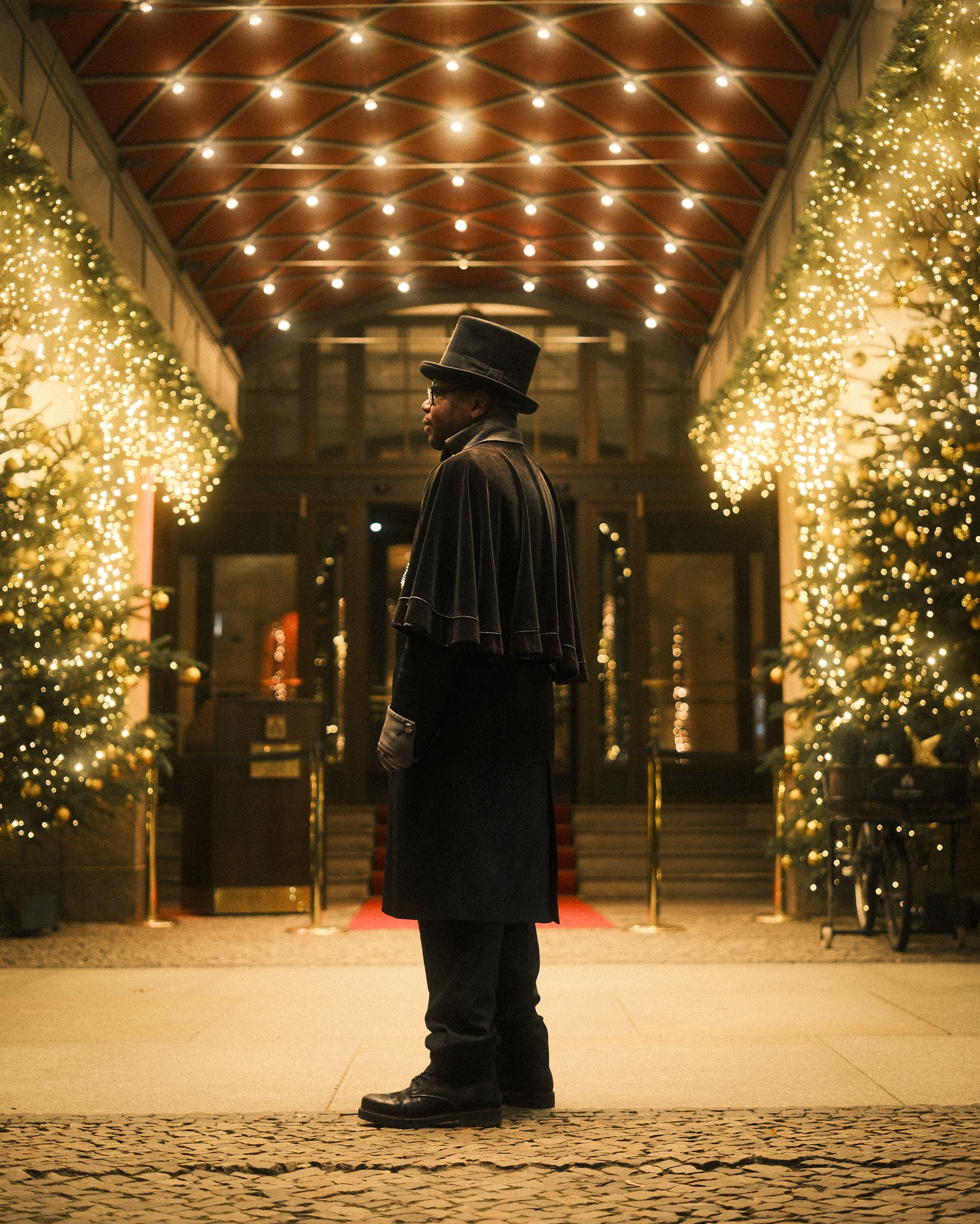A man in vintage attire stands under festive lights and trees in Berlin during Christmas season.