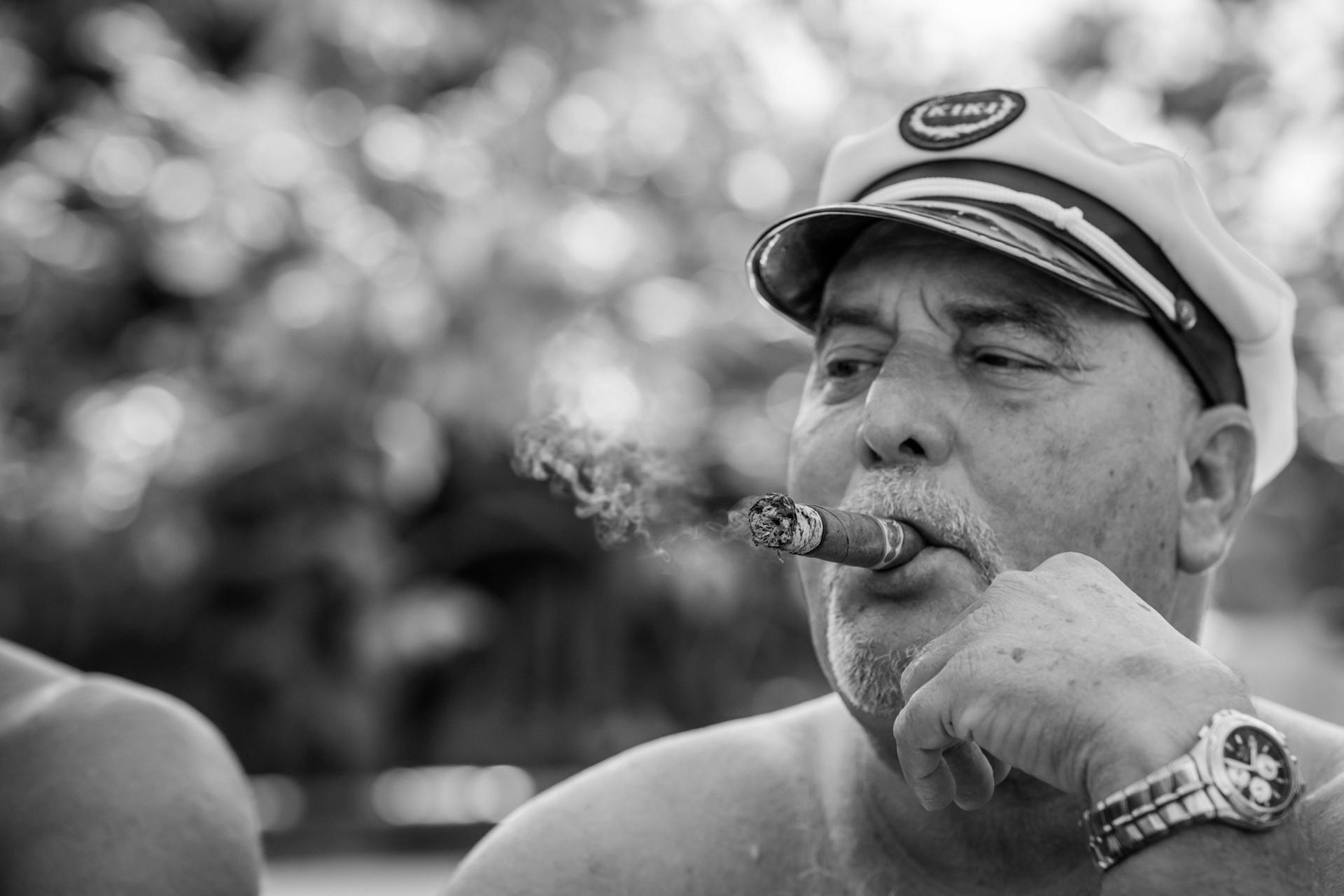 Black and white portrait of a senior man smoking a cigar outdoors, wearing a captain's hat.