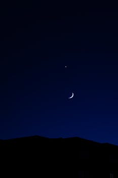A serene night sky with a crescent moon and visible star above silhouetted rooftops.