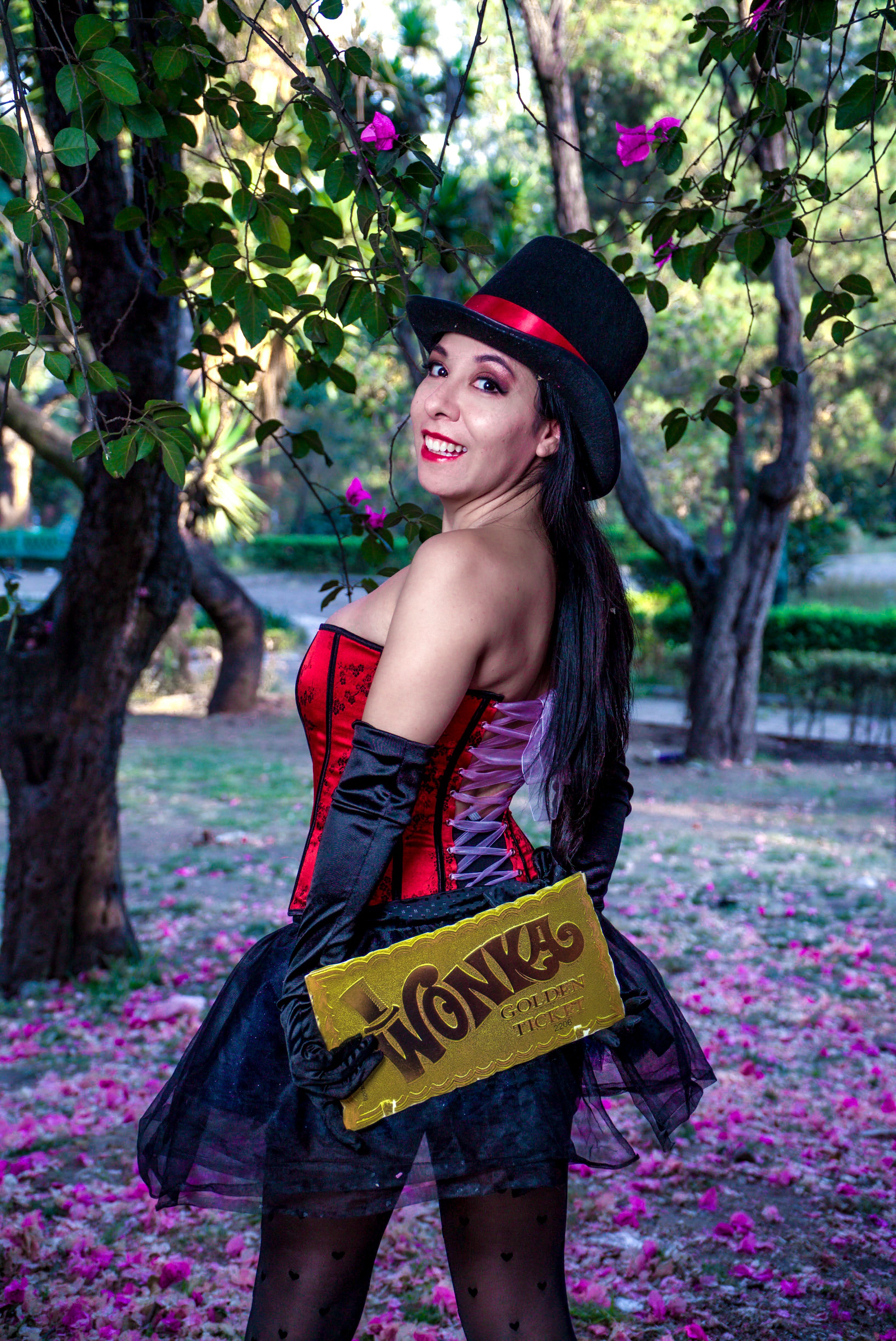 Woman in a whimsical costume holding a golden ticket outdoors, surrounded by trees and flowers.