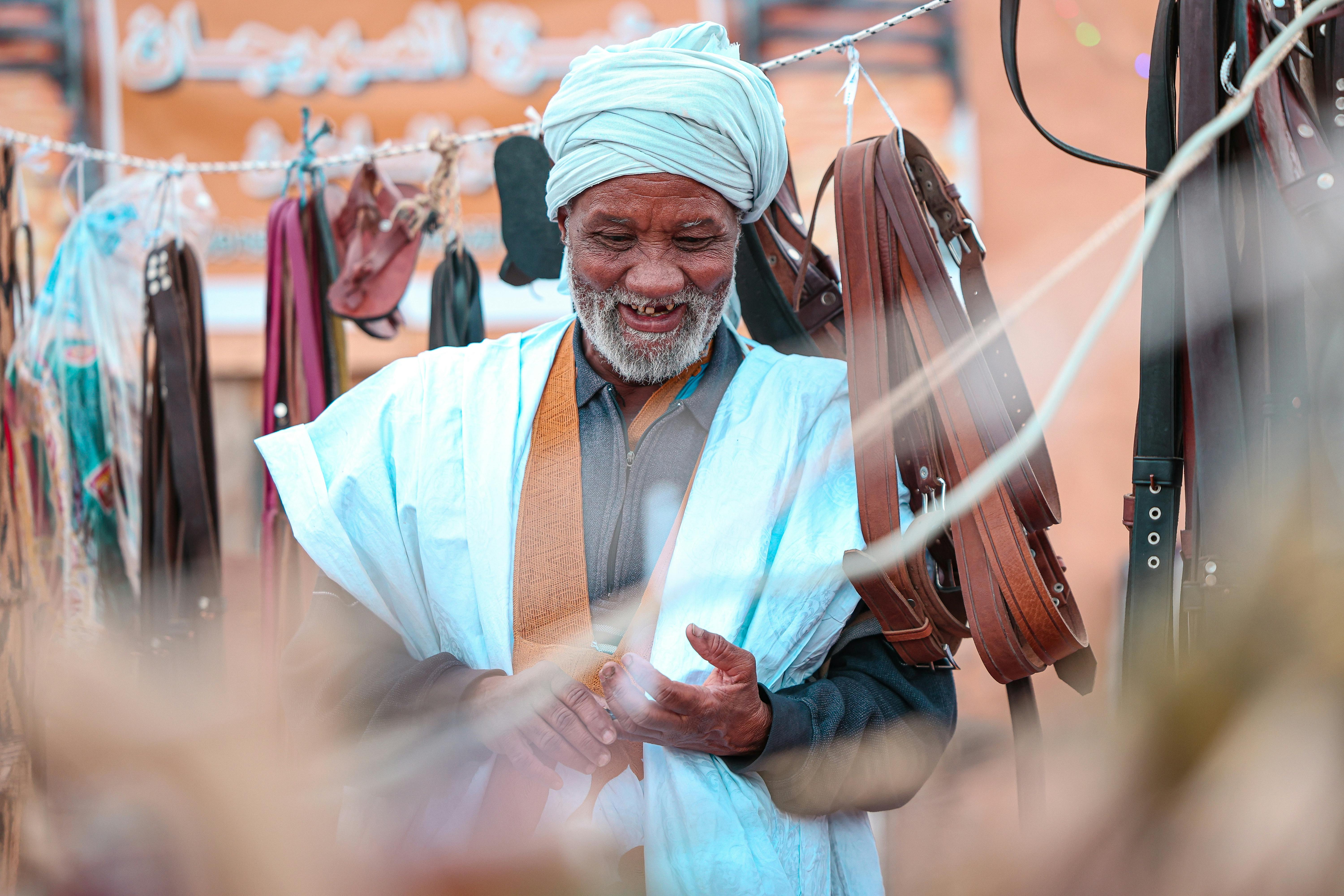 Smiling elderly man in traditional attire standing among leather goods at an outdoor market.