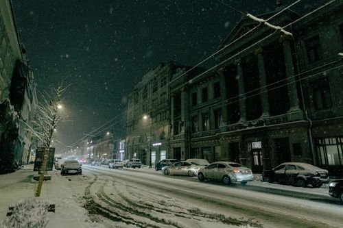 Free A tranquil urban street at night, covered in snow with parked cars and historic buildings. Stock Photo