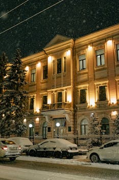 A historic building illuminated at night under falling snow, with cars parked nearby.