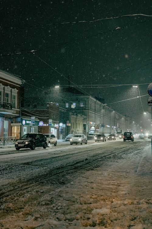 Free Cars driving on a snowy city street at night, creating a wintery urban atmosphere. Stock Photo