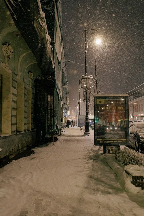Free A nighttime urban street covered in snow, featuring streetlights and parked cars. Stock Photo