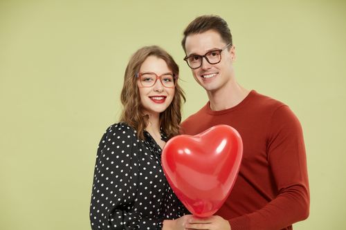 Free Smiling couple wearing glasses holding a red heart-shaped balloon against a soft pastel background. Stock Photo