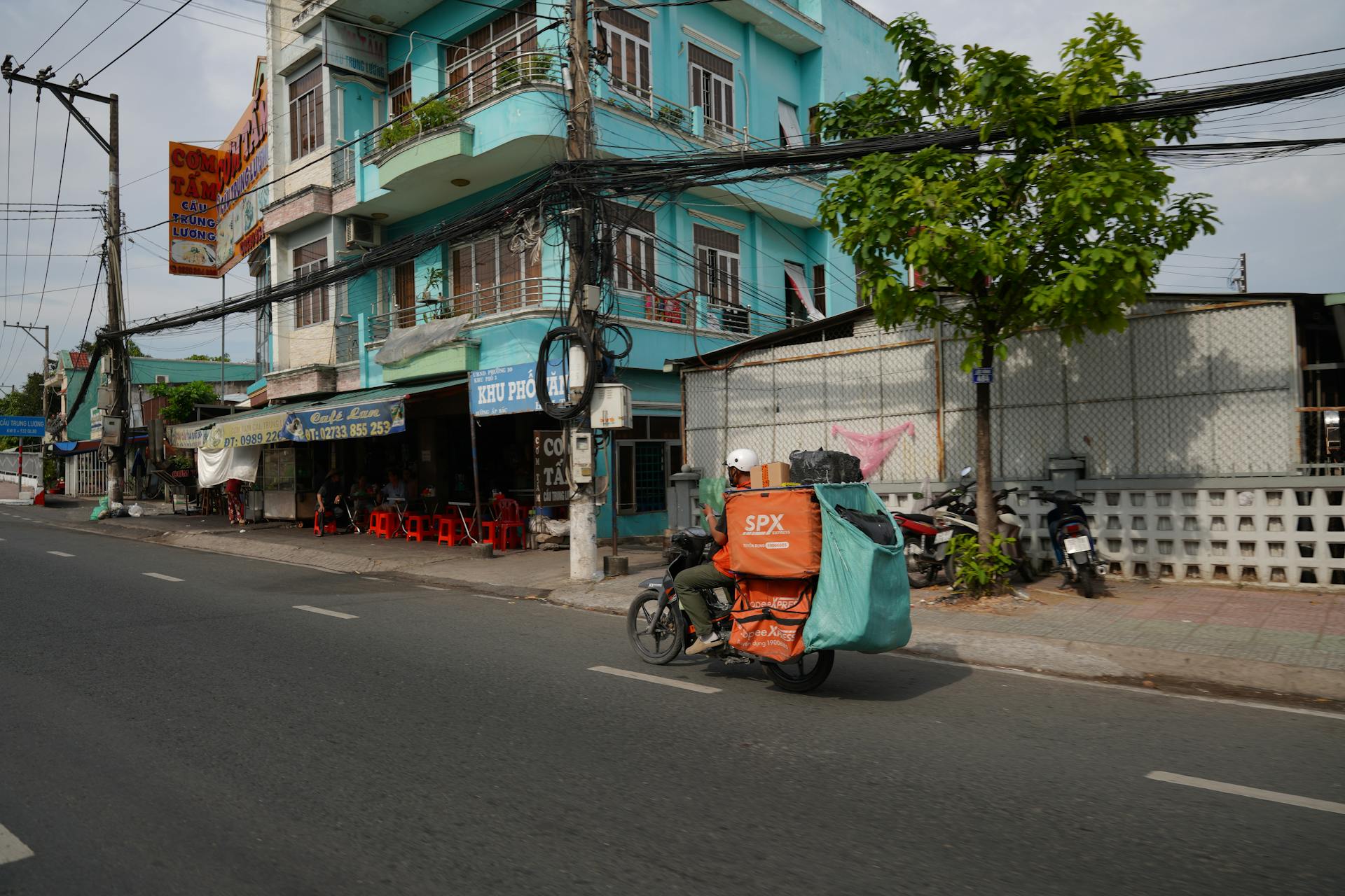 A delivery driver rides a motorcycle past colorful buildings on a sunny urban street.