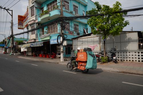 Free A delivery driver rides a motorcycle past colorful buildings on a sunny urban street. Stock Photo