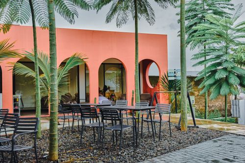 Free Outdoor seating at a vibrant cafe with tropical palm trees, featuring a coral-colored facade with arched windows. Stock Photo