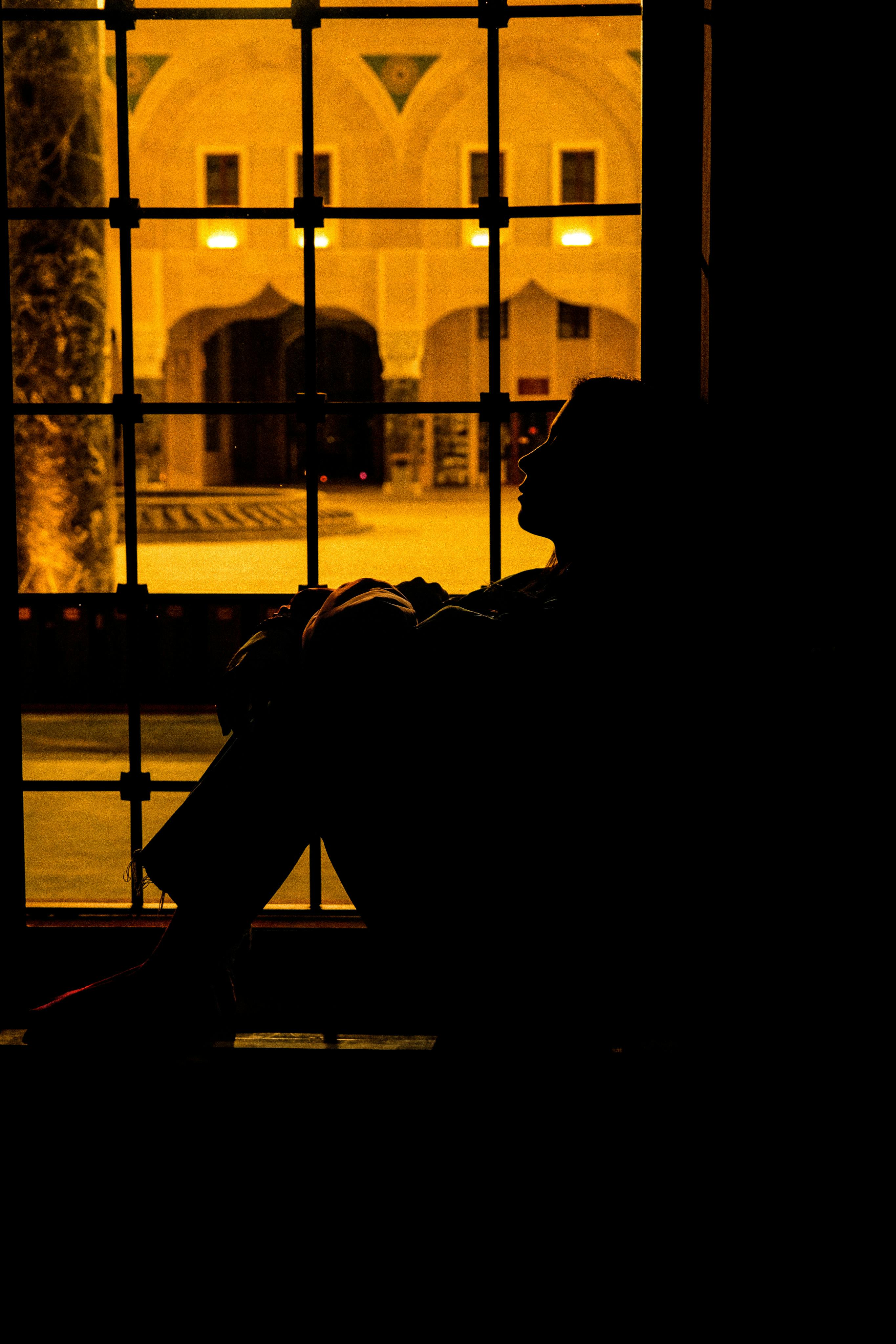 Kostenlos Die Silhouette einer Person, die an einem Fenster sitzt, mit einem beleuchteten historischen Gebäude im Hintergrund. Stock-Foto