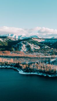 A picturesque view of snowy mountains, a serene lake, and autumn-colored trees in winter.