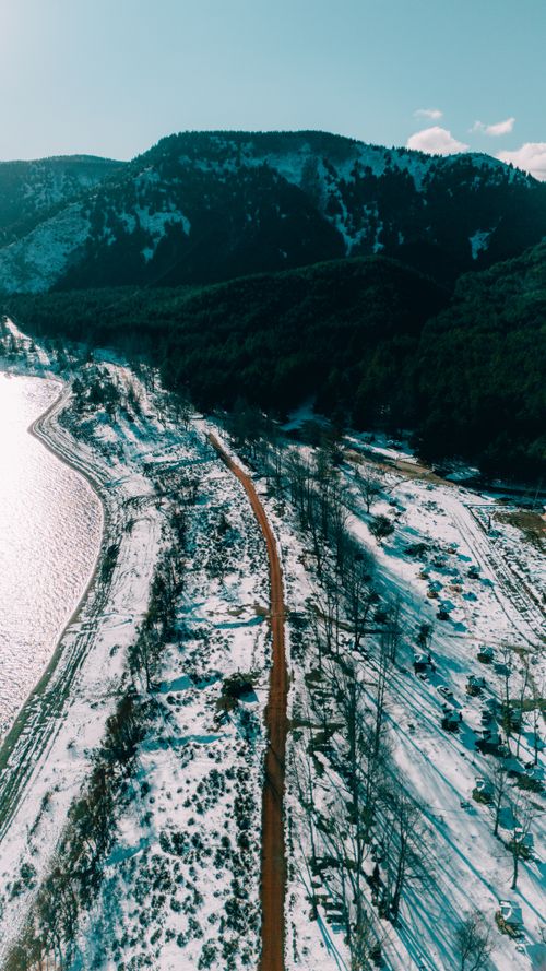 Free Stunning aerial view of a snowy landscape with a winding path beside a mountain lake. Stock Photo