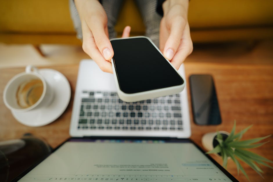 Hands holding smartphone over laptop in cozy home office setup with coffee and plant.