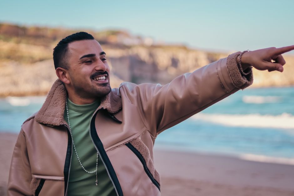 Smiling man wearing a stylish jacket pointing towards the ocean on a sunny beach day.