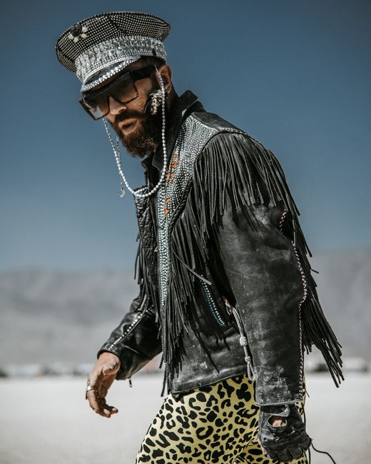 Fashionable man with fringe jacket and sunglasses posing outdoors in a desert environment.