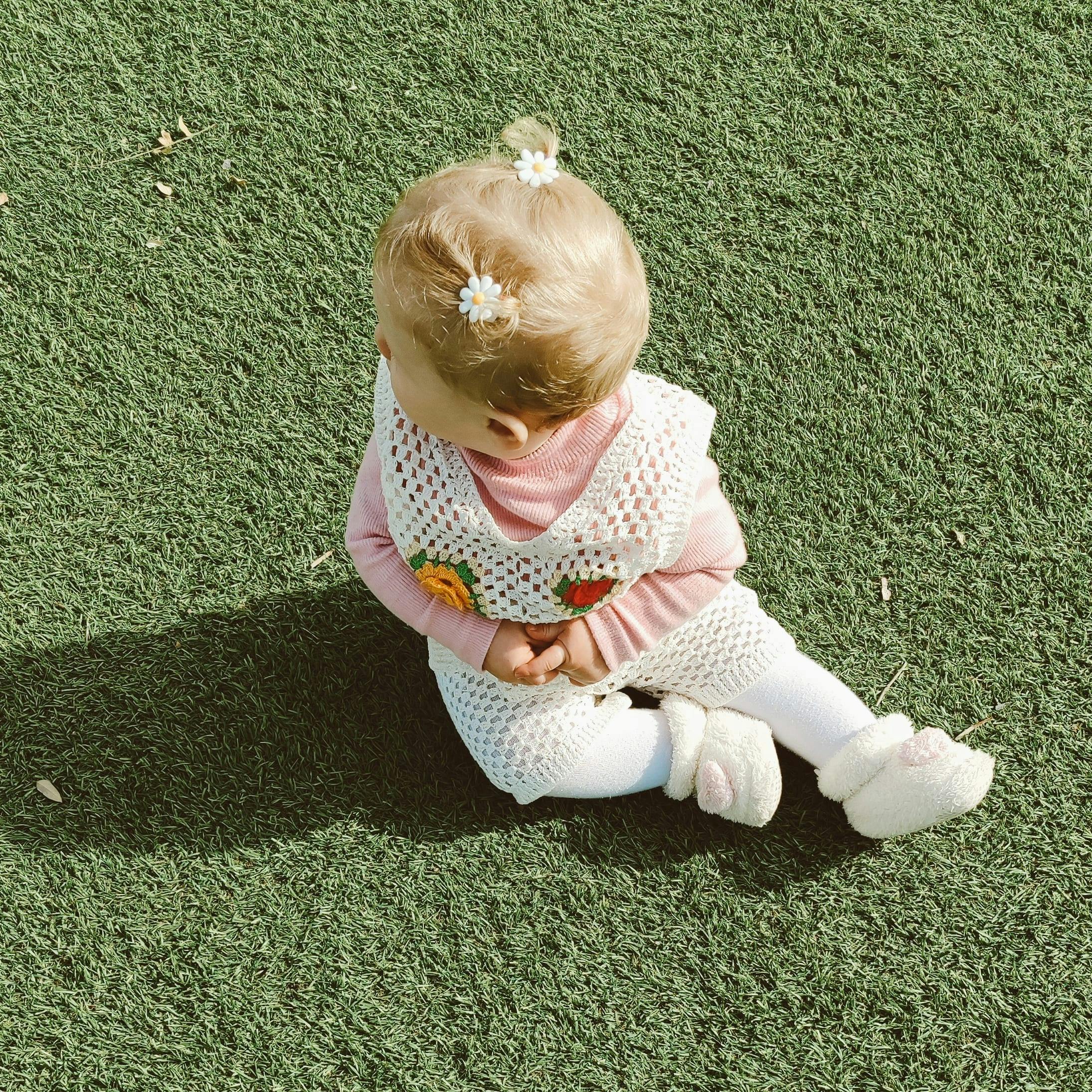 A cute baby girl sitting on lush grass wearing a handmade crochet outfit and flower hair accessory.