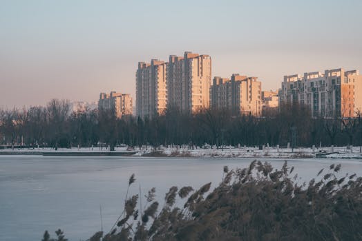 A serene winter urban landscape featuring buildings reflected in a frozen lake at sunrise.