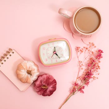 A pink-themed flat lay featuring a retro clock, coffee mug, notebook, and decorative pumpkins.