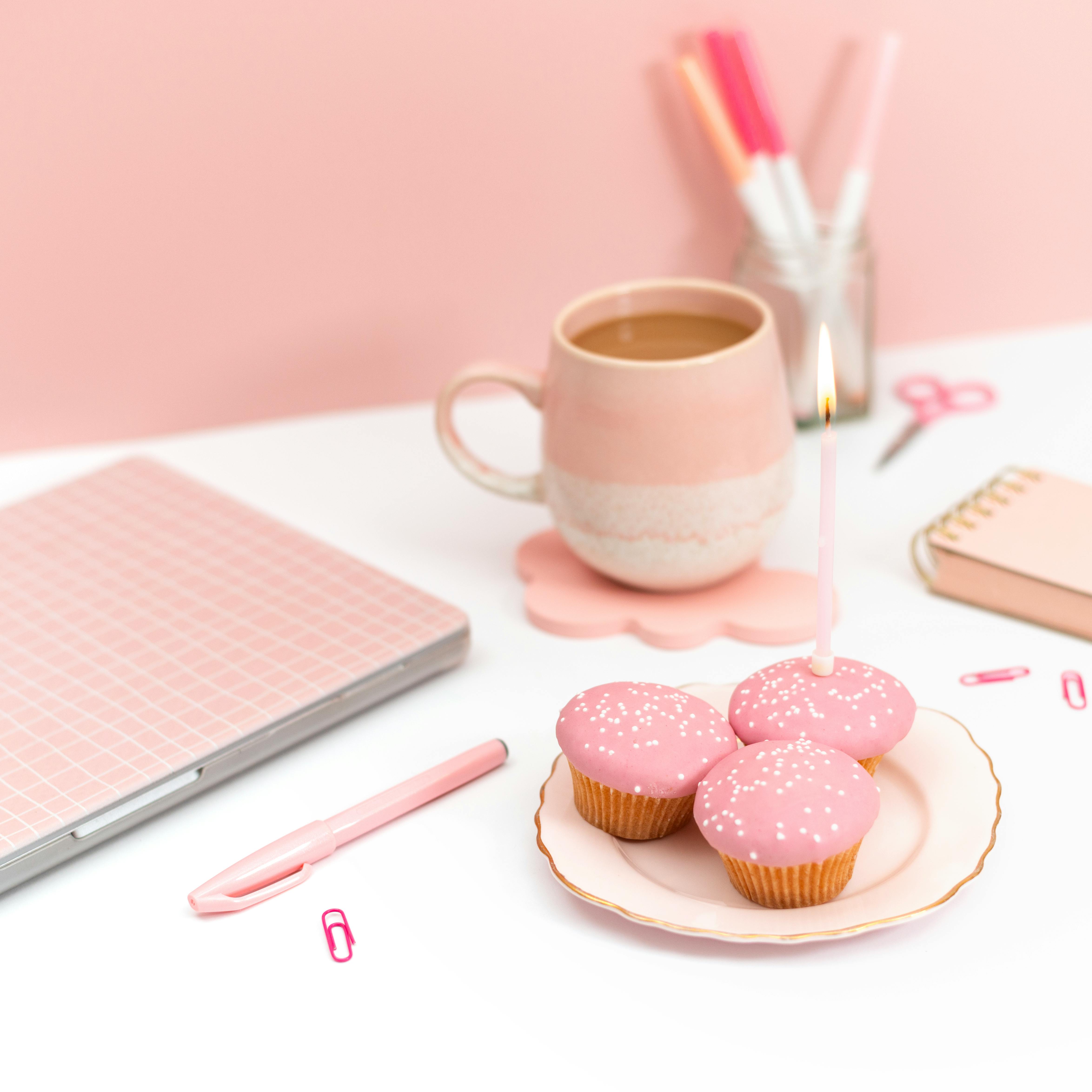 Elegant pink-themed workspace featuring a laptop, cupcakes with candles, and coffee mug.