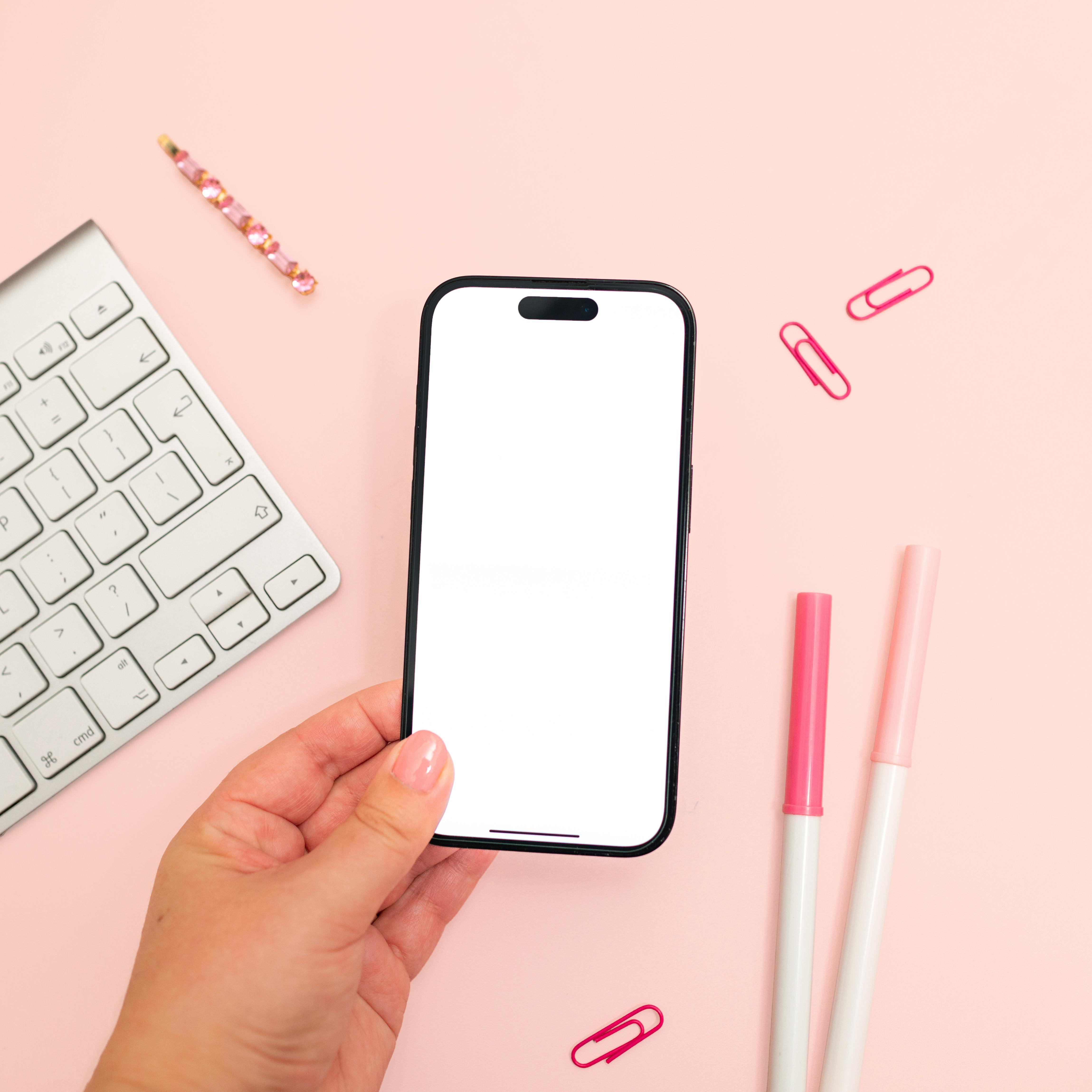 Free A minimalistic pink-themed workspace showcasing a smartphone mockup held in hand. Stock Photo