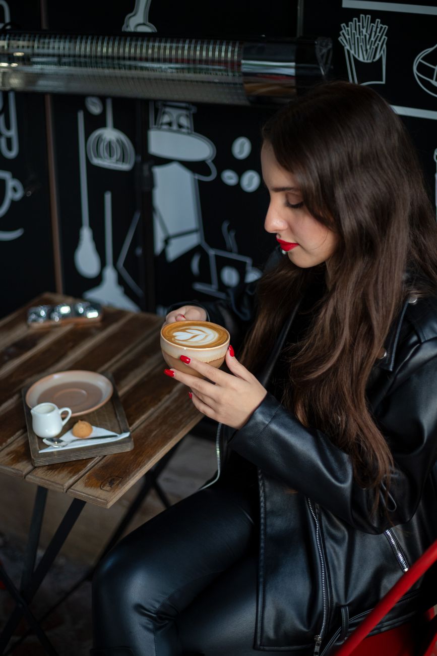 Stylish woman in leather jacket savoring a cappuccino at a modern cafe.