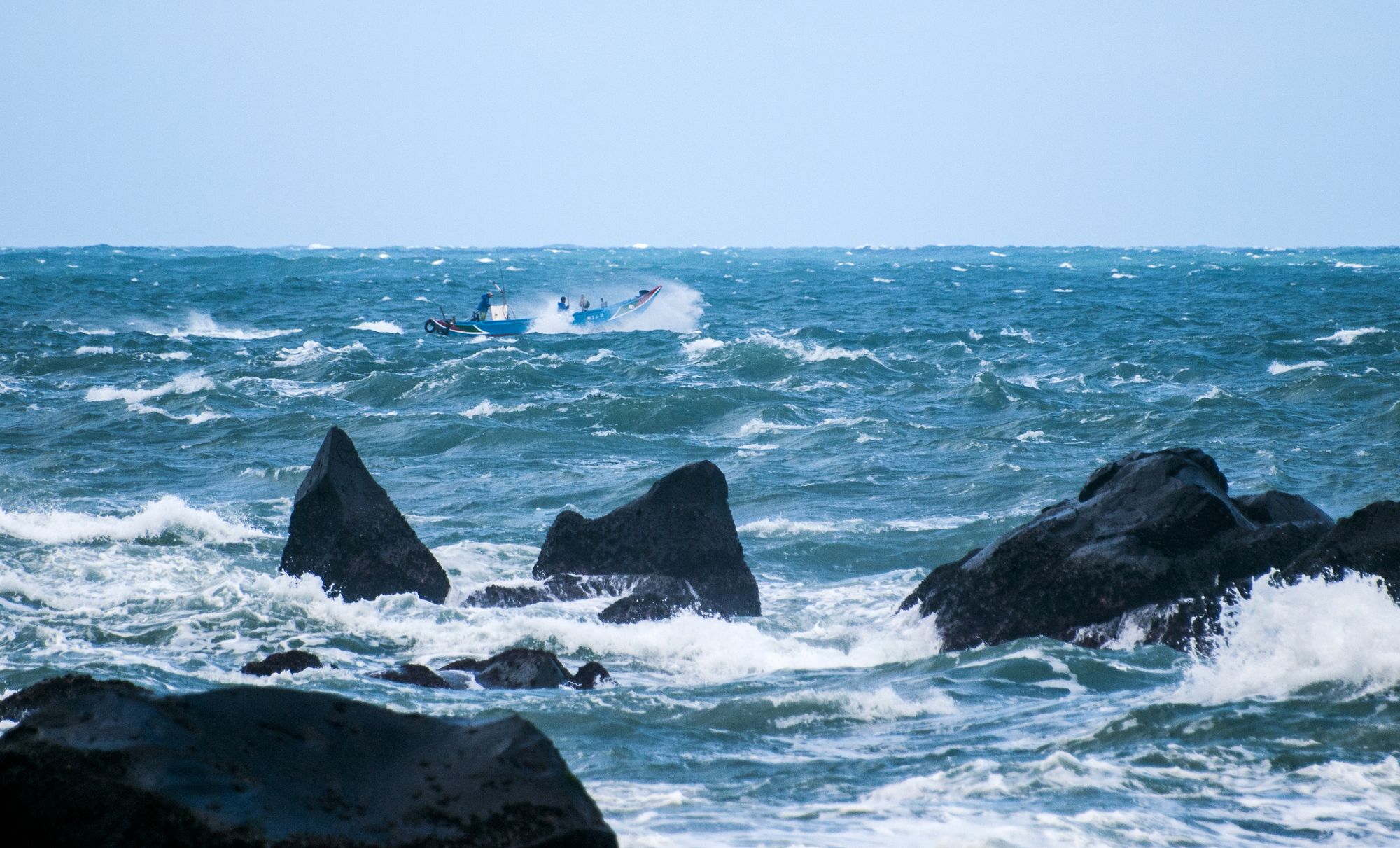 A fishing boat navigates through rough seas near rocky shores under a bright day sky.