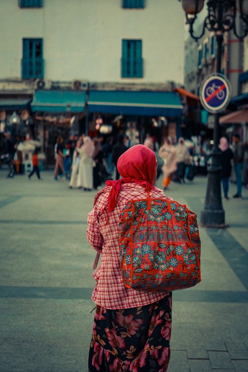 Free A woman wearing a floral backpack and red scarf in a bustling city market square, capturing everyday life. Stock Photo