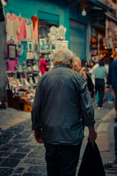 Free Back view of an elderly man walking in a vibrant outdoor market street filled with shops and people. Stock Photo
