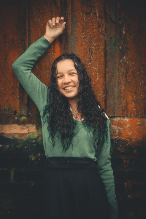 Photo of Smiling Girl in Green Long-sleeved Shirt Leaning on Wall Posing with Her Hand Up