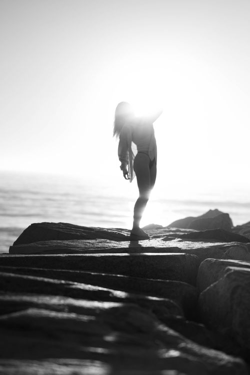 Free Artistic black and white photo of woman in silhouette standing on rocks by the sea at sunset in California. Stock Photo