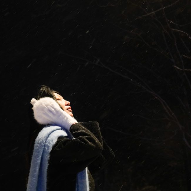 Adult woman with earmuffs enjoying falling snow at night outdoors.