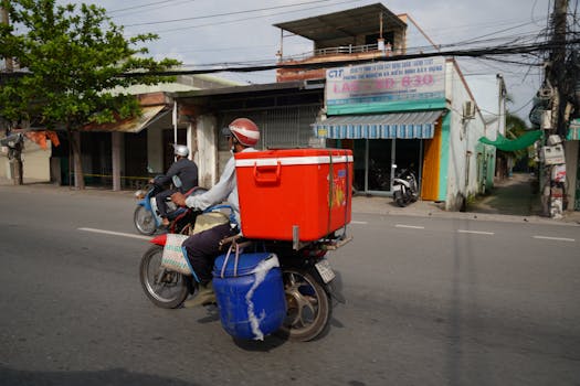 A person riding a delivery motorcycle carrying a red container on an urban street.
