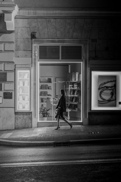Free A black and white photo of a woman walking past a brightly lit storefront at night. Stock Photo