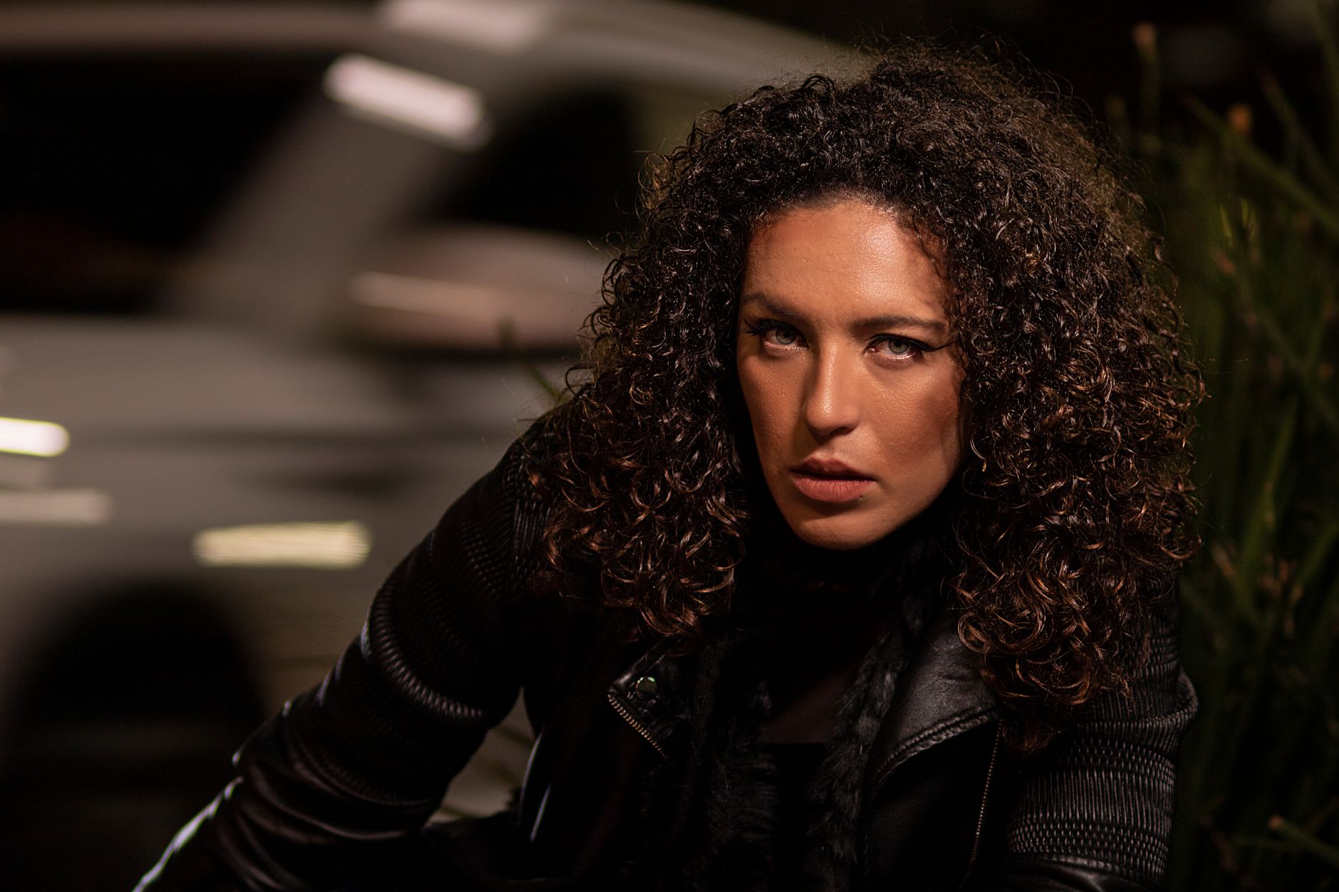 Close-up portrait of a woman with curly hair in a leather jacket under night lighting.