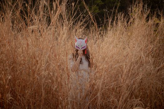 A person wearing a fox mask stands hidden in a field of tall grass, creating an enigmatic scene.