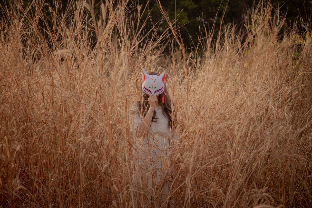 a person wearing a fox mask stands hidden in a field of tall grass cre