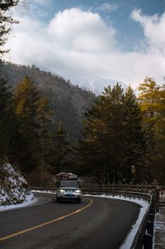 A car drives on a snow-lined mountain road surrounded by tall pine trees and cloudy skies.