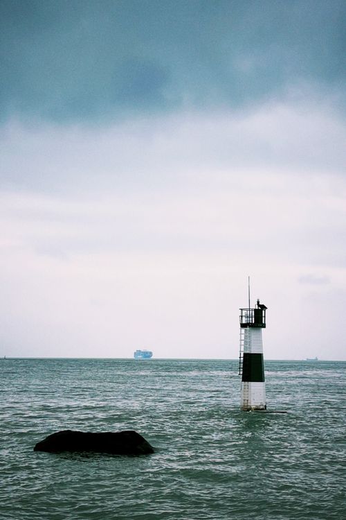 Free Lighthouse standing alone in the ocean with distant ship under cloudy skies, depicting solitude and navigation. Stock Photo