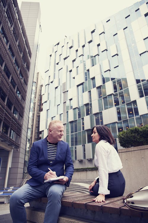 Man and Woman Sitting on Wooden Bench Outside Buildings