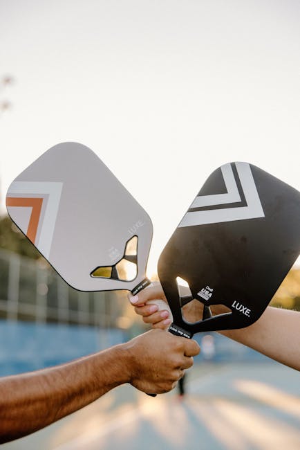 Two hands holding pickleball paddles outdoors during sunset.