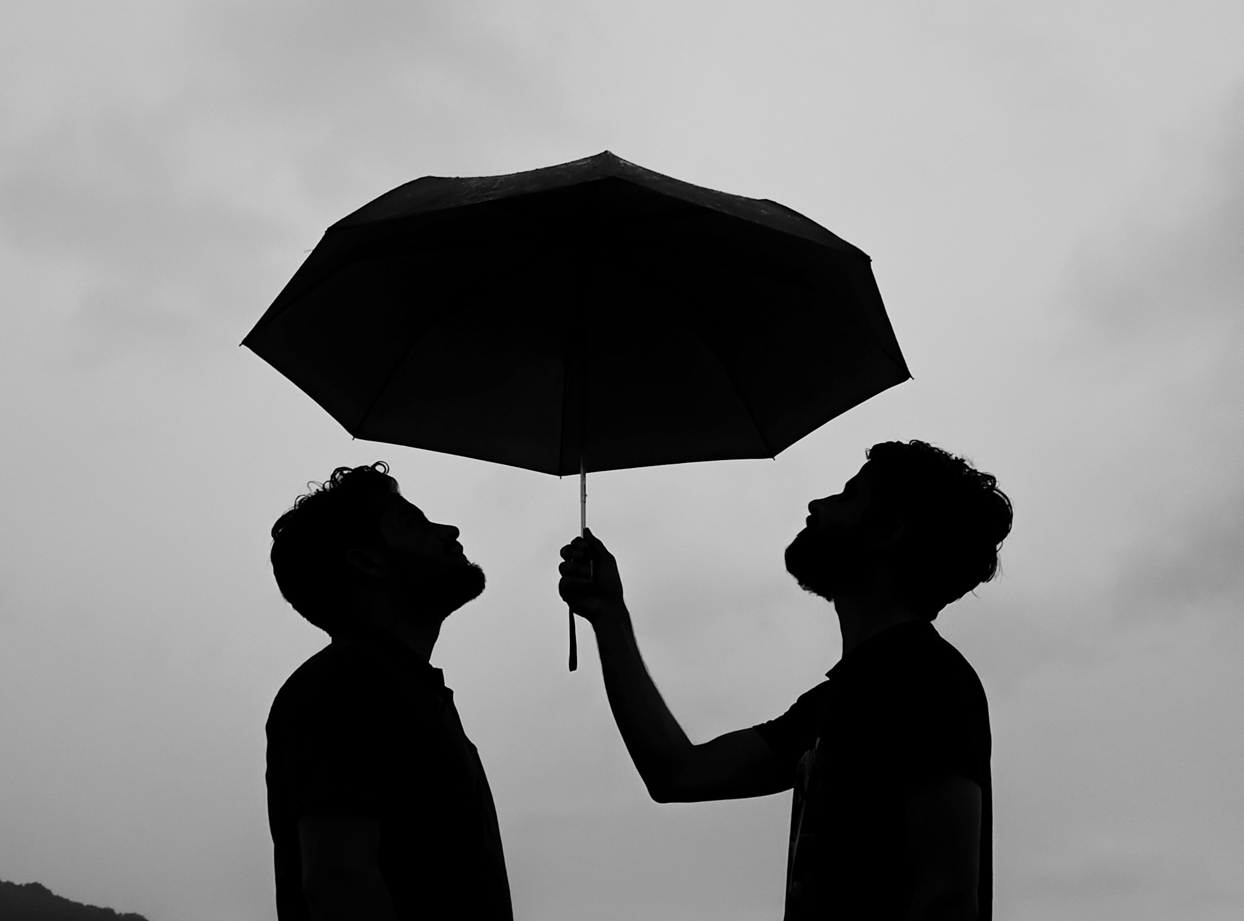 Free Two silhouetted individuals stand beneath an umbrella against a cloudy sky in monochrome. Stock Photo