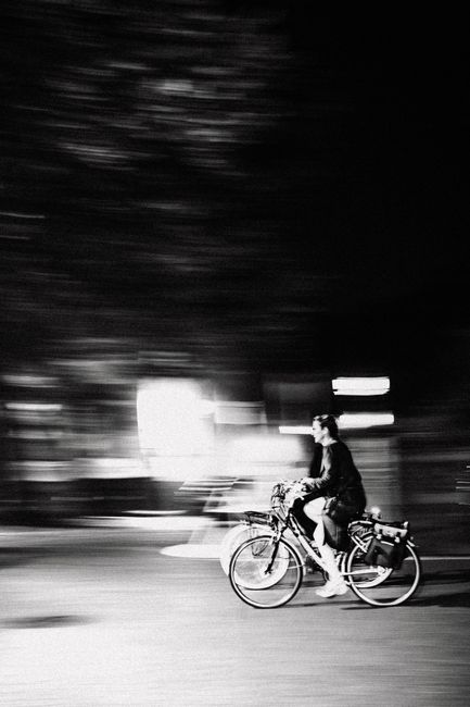 A blurred black and white photo of a cyclist riding through Bruges at night.